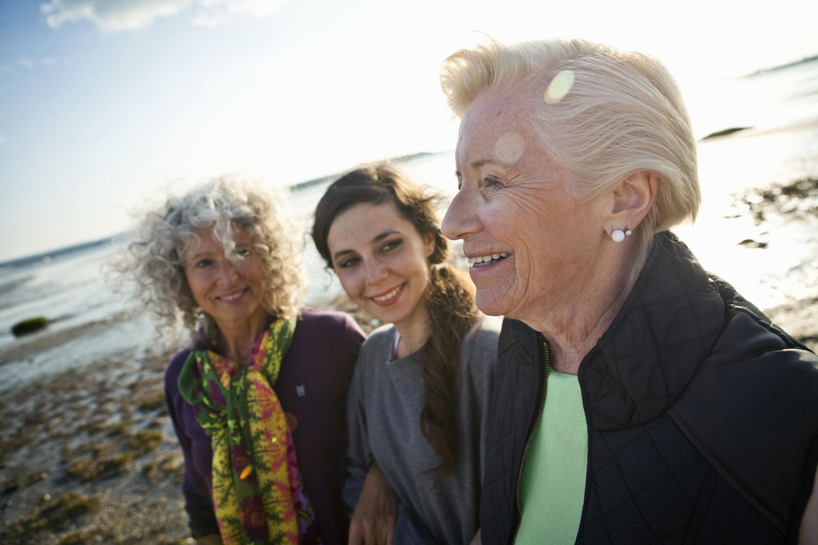 Three generations of women: grandmother, mother, and daughter smiling together while walking on a sunny beach.