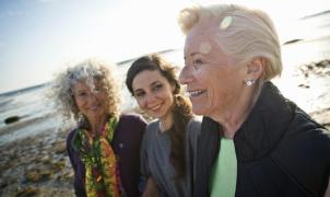 Three generations of women: grandmother, mother, and daughter smiling together while walking on a sunny beach.